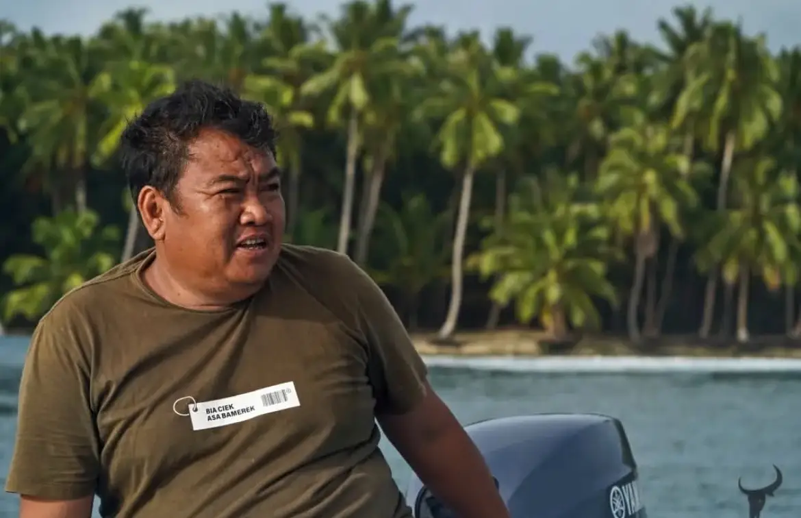 Captain Pak Iwan at the tiller of a tender with a palm-covered shoreline behind