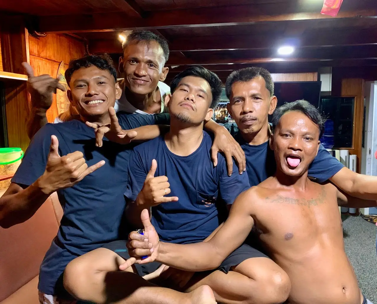 Five smiling crew members giving shaka signs in the boat's saloon