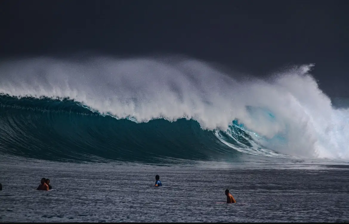 A heavy thick-lipped wave breaking under a dark storm sky with surfers in the channel