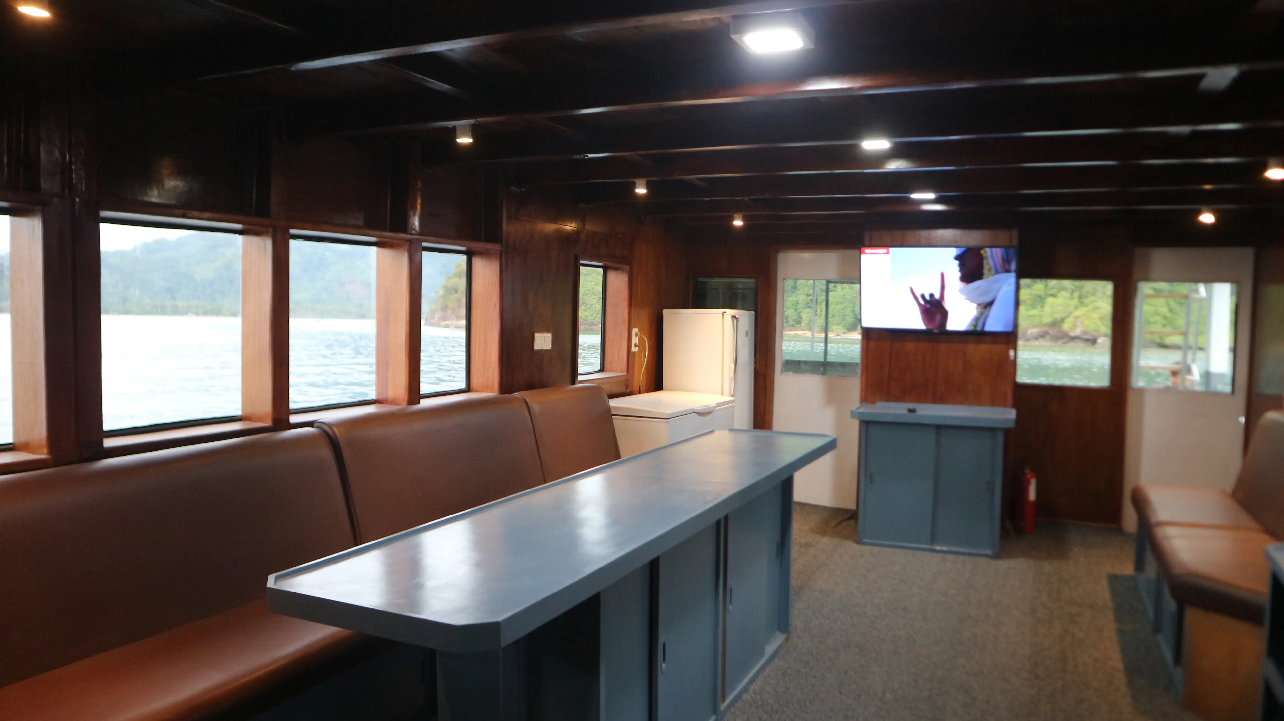 Main saloon with long padded bench seating, central table and windows onto the shoreline