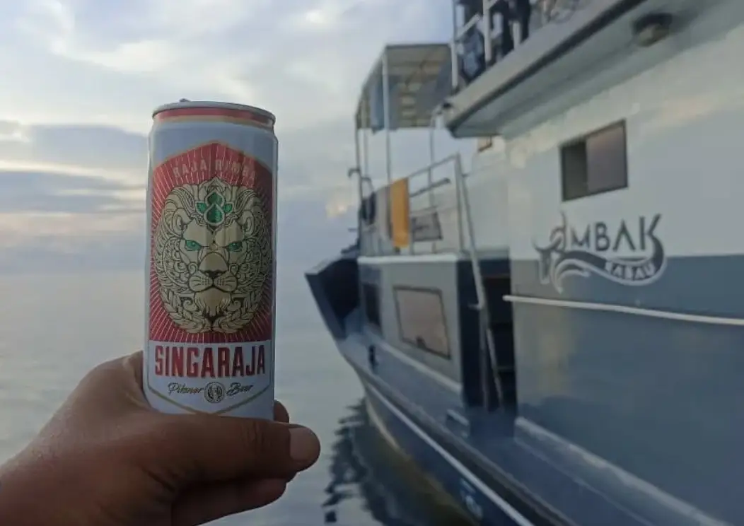 A cold beer held up at sunset with the Ombak Kabau name graphic behind